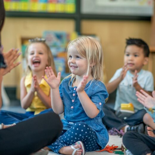 Children sitting on the floor in a classroom, smiling and clapping along with an adult.